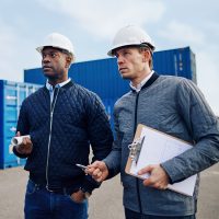 Two engineers wearing hardhats and discussing logistics together while standing in a freight yard full of shipping containers